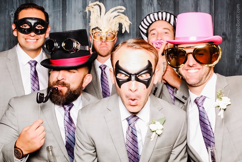 Six men in suits posing playfully with various props, including masks and oversized sunglasses, in front of a Blackwood backdrop at Las Positas Vineyard, Livermore.