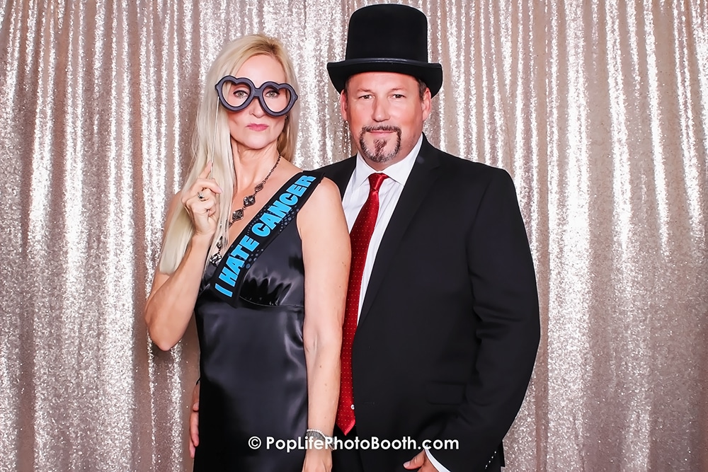 Couple posing in a photo booth with a silver sequins backdrop, woman wearing oversized glasses and a sash that reads "I HATE CANCER," and man in a black suit and top hat, reflecting a fun event atmosphere.