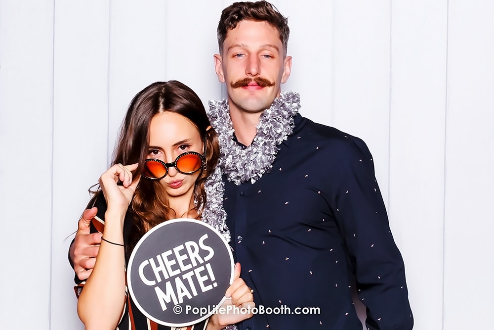 Couple posing playfully in a photo booth with a "Cheers Mate!" sign, featuring festive attire and accessories, capturing a fun moment at an event in Livermore, California.