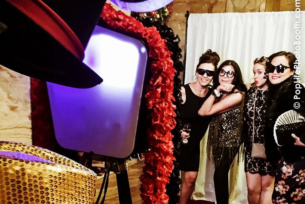 Group of four women in glamorous attire posing playfully in front of a photo booth with a stylish backdrop, capturing the festive atmosphere of an event at a San Francisco venue.