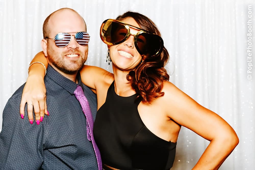 Couple posing in photo booth at The Press Club, San Francisco, wearing oversized sunglasses and smiling, with a sparkling backdrop.