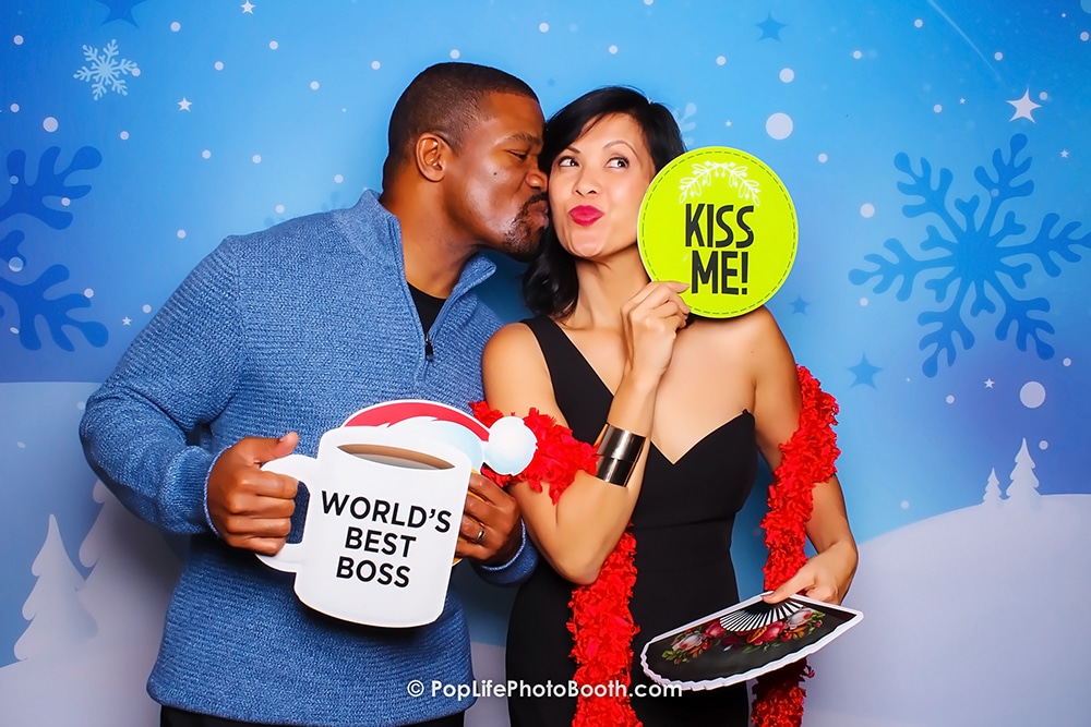 Couple sharing a kiss in a photo booth with a Winter Wonderland backdrop, holding props including a "World's Best Boss" mug and a "Kiss Me!" sign, showcasing a fun and festive atmosphere.