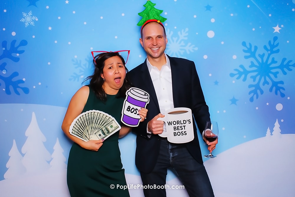 Couple posing in a photo booth with festive winter backdrop, holding props including money, coffee cup labeled "BOSS," and mug stating "WORLD'S BEST BOSS."