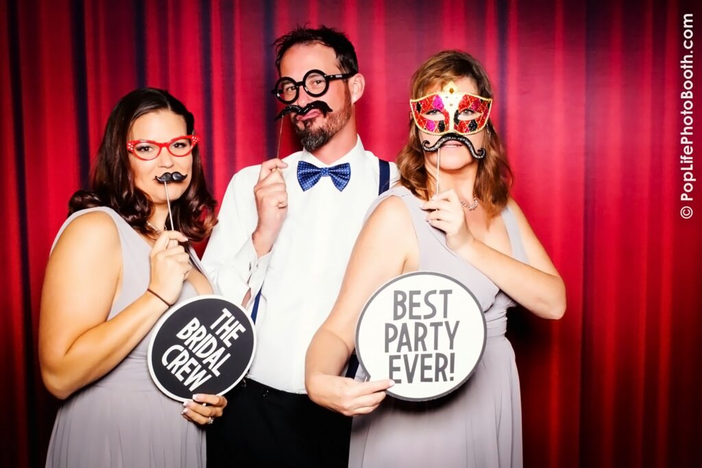 Three people posing in a photo booth with playful props, including a mustache and masks, in front of a vibrant red backdrop, highlighting a fun party atmosphere at an event.