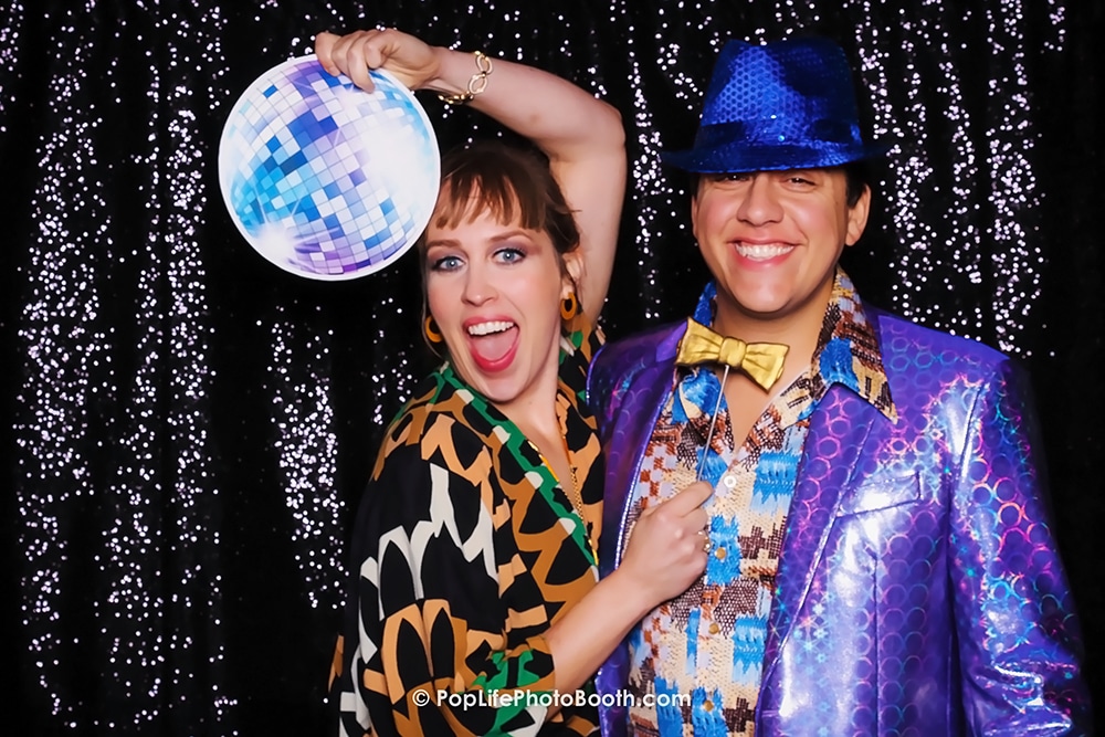 Couple posing in a photo booth with a disco ball, showcasing vibrant 70s-style outfits against a sparkling black backdrop, representing fun at The Press Club venue in San Francisco.