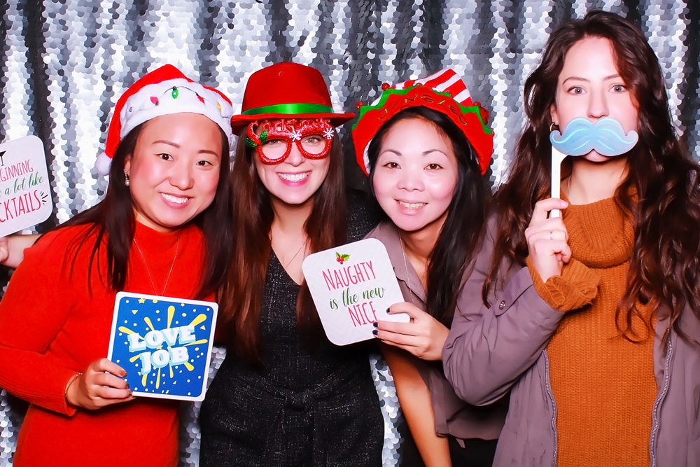 Four women in festive attire posing with playful props in front of a shimmering silver backdrop, celebrating at a holiday event.