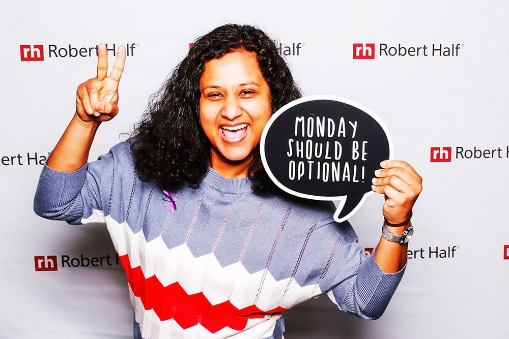 Smiling woman in a photo booth holding a sign that reads "MONDAY SHOULD BE OPTIONAL!" with Robert Half branding in the background, representing a fun corporate event atmosphere.