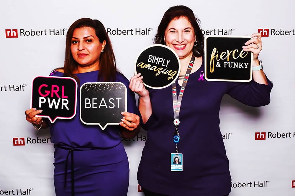 Two women at a corporate event holding colorful signs that read "GRL PWR," "BEAST," "SIMPLY amazing," and "fierce & FUNKY," set against a branded backdrop featuring Robert Half logo, showcasing a fun and engaging atmosphere for event attendees.
