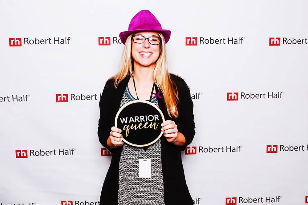 Woman wearing a purple hat and glasses holding a "WARRIOR queen" sign in front of a branded backdrop for Robert Half, showcasing a fun moment at a corporate event photo booth.