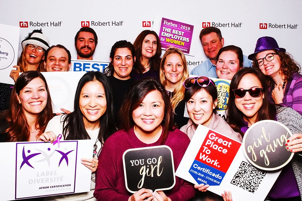 Group of diverse individuals smiling in a photo booth, holding signs promoting workplace diversity and recognition, with a backdrop featuring the Robert Half logo, reflecting a corporate event atmosphere.