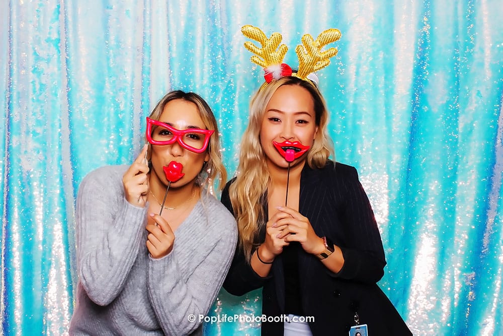 Two women posing in a photo booth with festive props, including oversized glasses and red lips, against a shimmering blue sequined backdrop, capturing the fun atmosphere of a holiday party.