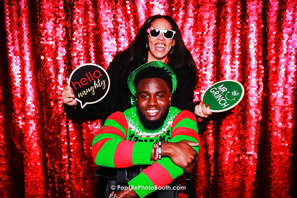 Couple posing in a photo booth with festive holiday attire, holding signs that read "hella naughty" and "Mr. Grinch," against a shimmering red sequin backdrop, capturing the joyful spirit of a holiday party.