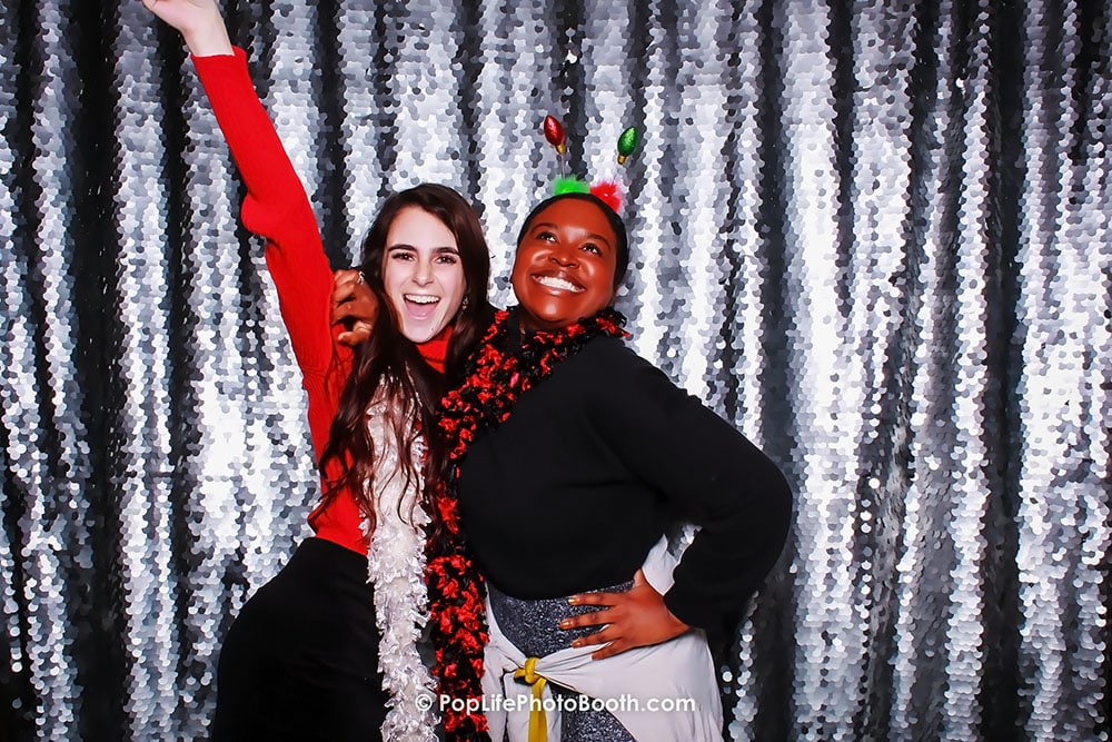 Two guests joyfully posing in a photo booth with a festive winter backdrop, wearing holiday attire and accessories, celebrating a holiday party.