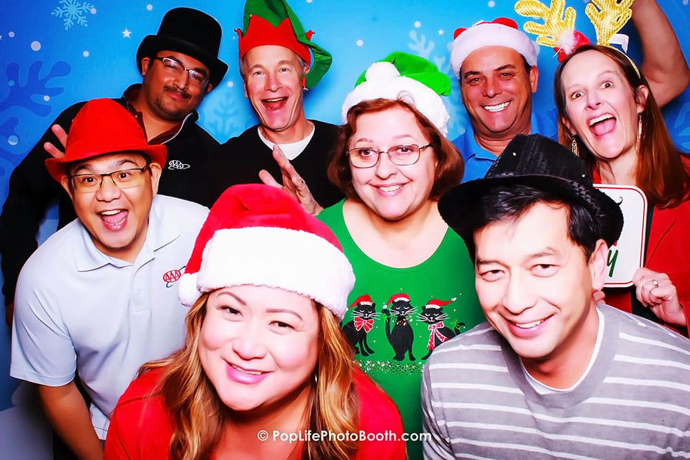 Group of festive guests in holiday attire, smiling with Santa hats and other holiday accessories, posing in front of a Winter Wonderland backdrop at a Photo Booth Holiday Party.