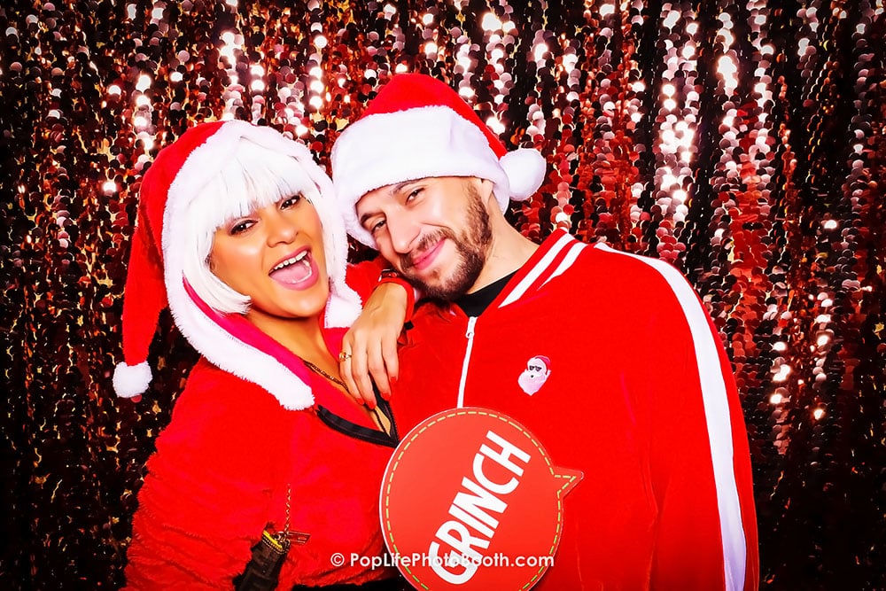 Couple in festive holiday attire with Santa hats posing joyfully in front of a shimmering sequin backdrop, holding a "GRINCH" sign at a Photo Booth Holiday Party.