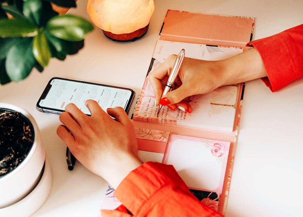 Hand writing a checklist in a planner with a smartphone displaying a calendar, surrounded by a plant and a lamp, emphasizing organization in event planning.