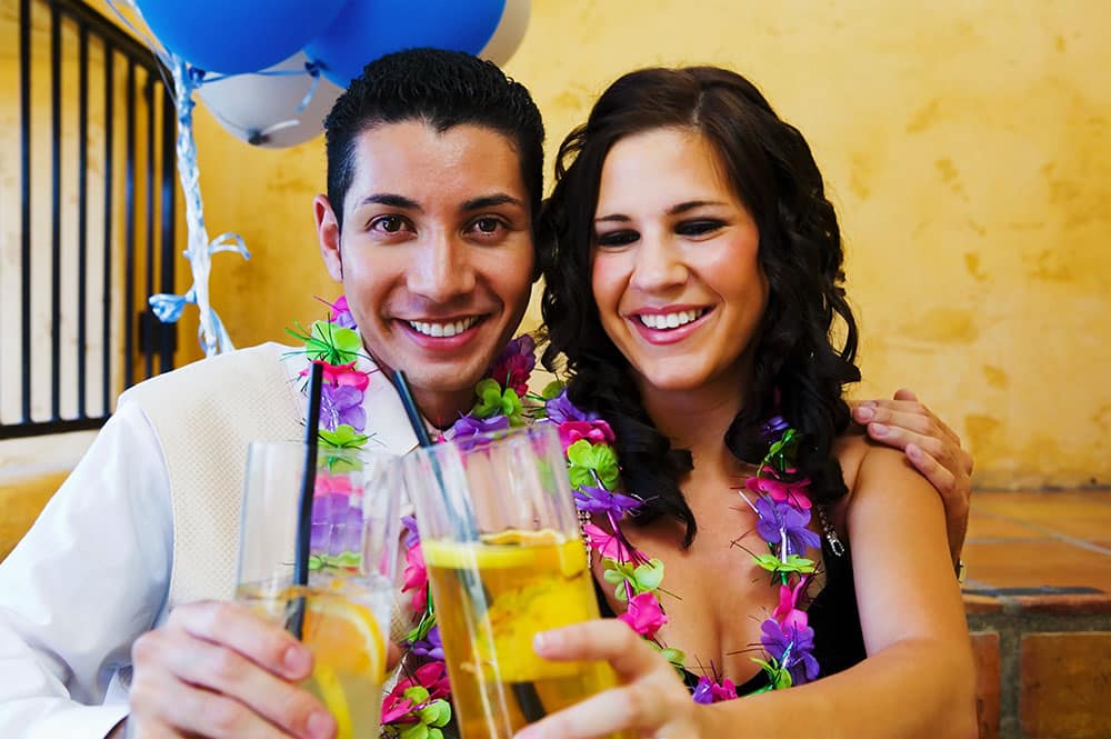 Couple celebrating at a party, wearing floral leis and holding cocktails, with festive decor in the background, representing a fun photo booth experience for event planning.