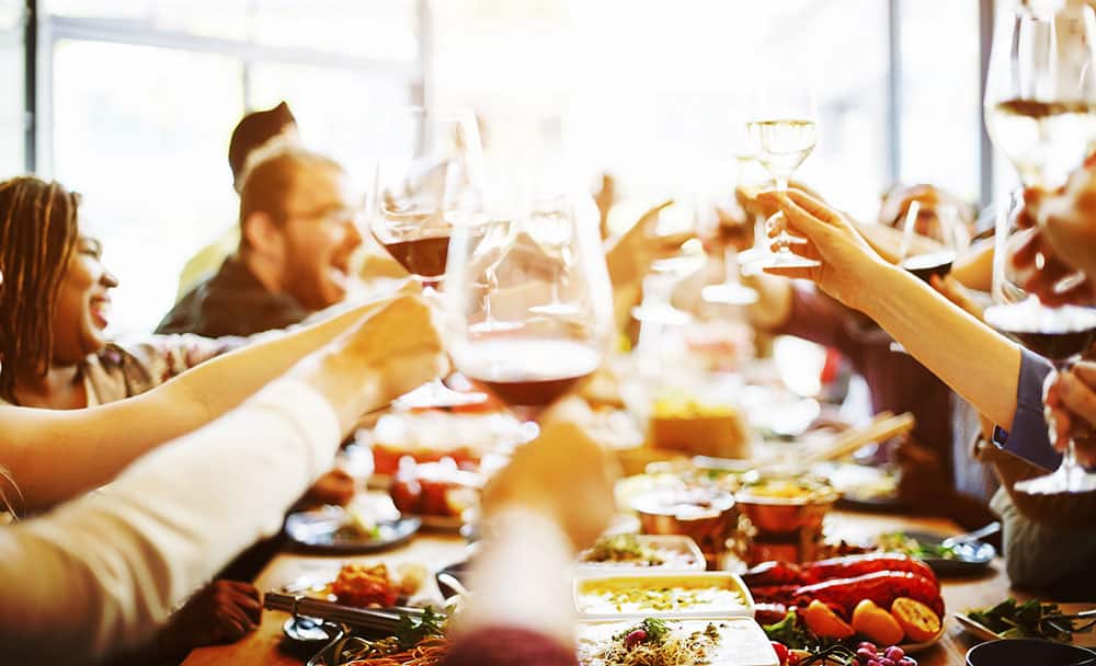 Group of people toasting with wine glasses at a festive dining table, celebrating during a holiday party with food and drinks.