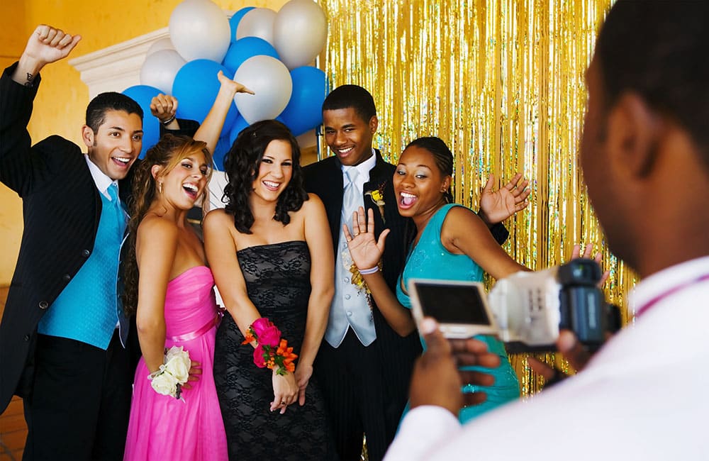Group of high school students posing joyfully in formal attire at a school dance photo booth, surrounded by balloons and a shiny backdrop, capturing fun memories.
