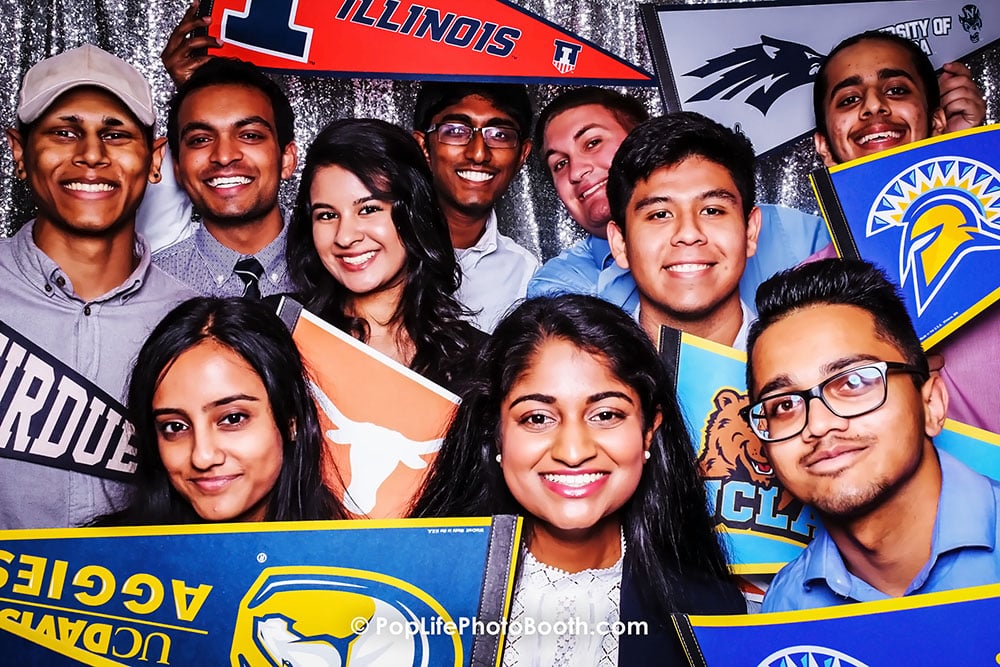Group of high school students joyfully posing in a photo booth, holding college banners, celebrating their upcoming graduations at a school event.