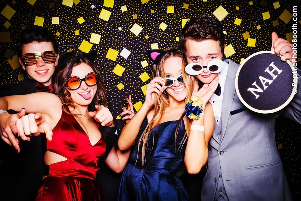 Students posing in a photo booth with fun props, wearing sunglasses and playful expressions, against a festive backdrop of gold and black decorations, capturing joyful moments at a school event.