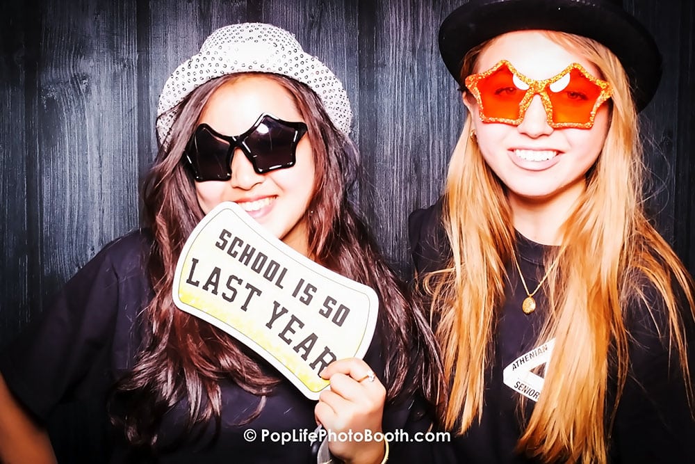 Two high school students wearing fun oversized sunglasses and hats, holding a sign that reads "SCHOOL IS SO LAST YEAR," celebrating at a photo booth during a school dance event.
