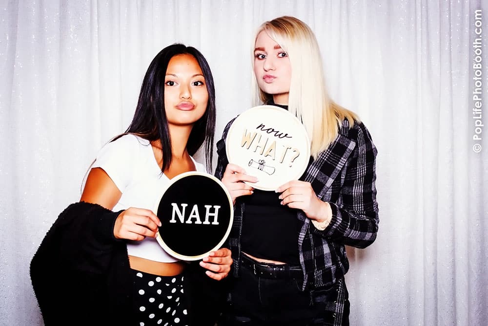 Two students posing in a photo booth holding signs that say "NAH" and "now WHAT?", celebrating at a school event.