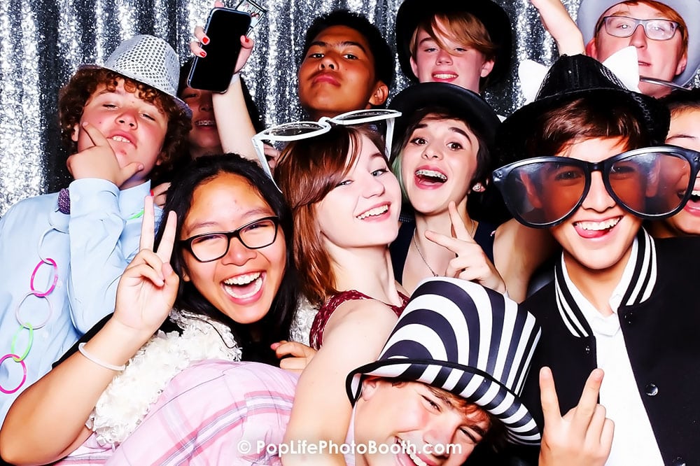 Group of high school students enjoying a photo booth at prom, wearing fun props like oversized glasses and hats, smiling and posing for the camera, with a sparkling backdrop.