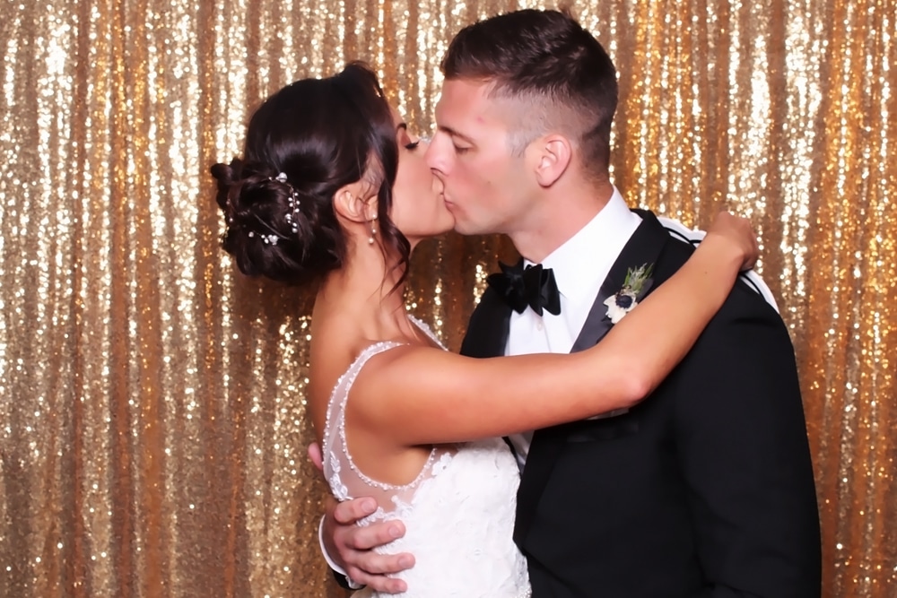Newlywed couple sharing their first kiss in a wedding photo booth with a sparkling gold backdrop at the San Ramon Marriott.