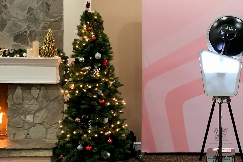 Christmas tree with lights and ornaments next to a stone fireplace, featuring a photo booth setup with a pink backdrop and lighting, in a festive event setting at Wedgewood Weddings in San Ramon.