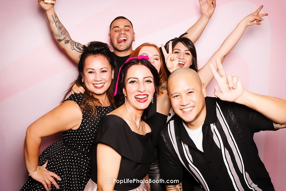 Group of six friends posing joyfully in front of a vintage blush backdrop at a holiday party, celebrating together with smiles and peace signs, highlighting the festive atmosphere at Wedgewood in San Ramon.