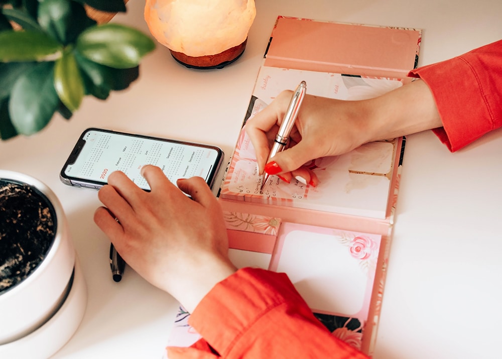 Woman writing a checklist in a planner, smartphone displaying a calendar, surrounded by a plant and a lamp, emphasizing last-minute event planning and task organization.