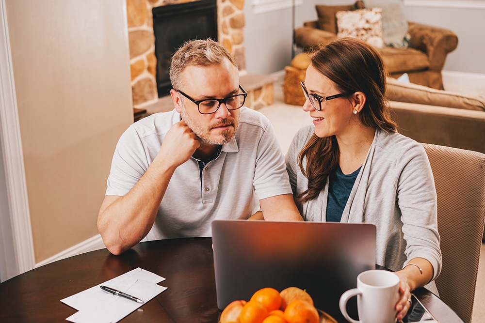 Couple collaborating on last-minute event planning at home, reviewing details on a laptop with notes and a cup of coffee nearby.