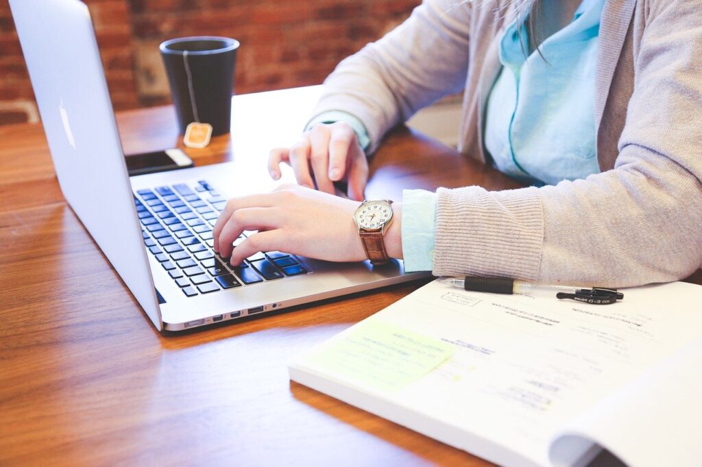 Person typing on laptop while planning event, with documents and coffee cup on wooden table, illustrating preparation for successful corporate or holiday event.
