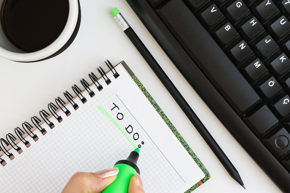 Woman highlighting "TO DO" on a checklist notebook, coffee cup and keyboard in the background, emphasizing organization for last-minute event planning.