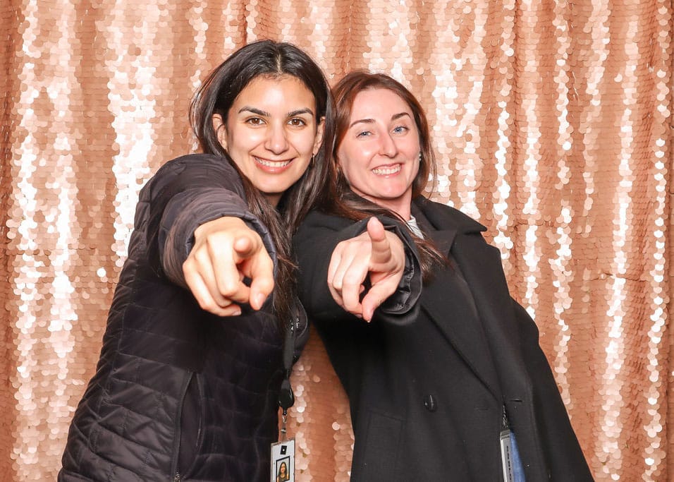 Two women posing playfully in a photo booth, pointing at the camera with smiles, against a shimmering rose gold backdrop, highlighting the fun and interactive atmosphere of corporate events.