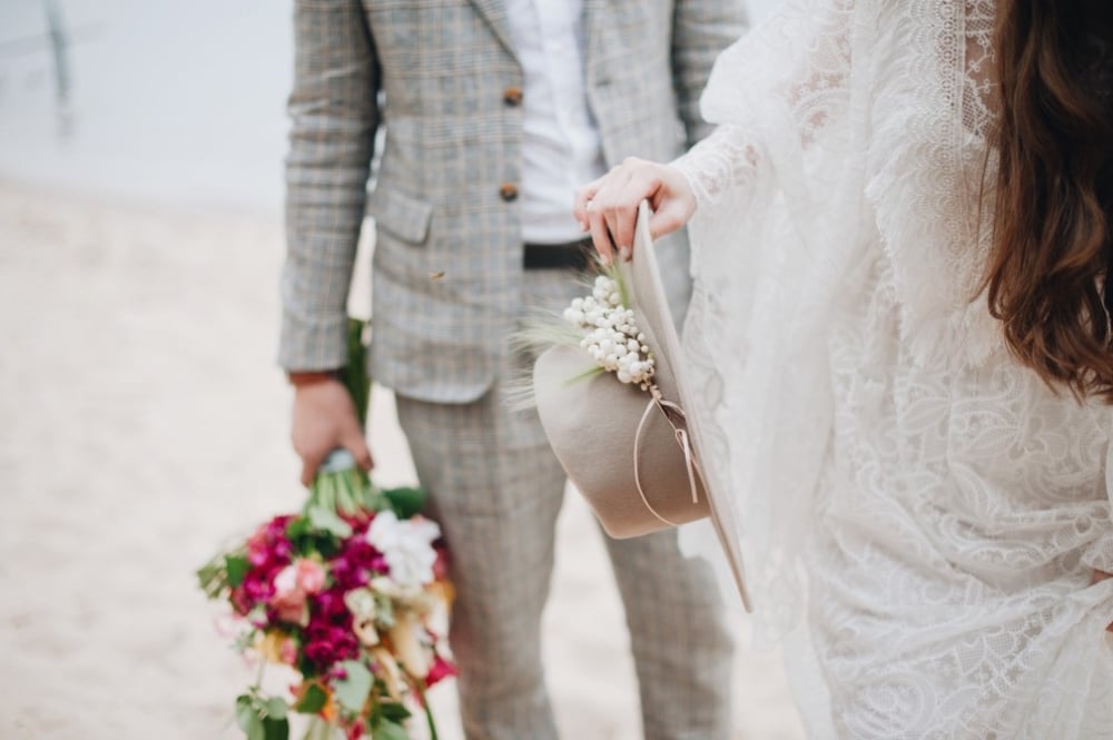 Bride in lace wedding dress holding a hat with floral accents and bouquet of vibrant flowers, standing beside groom in a checked suit, beach setting.