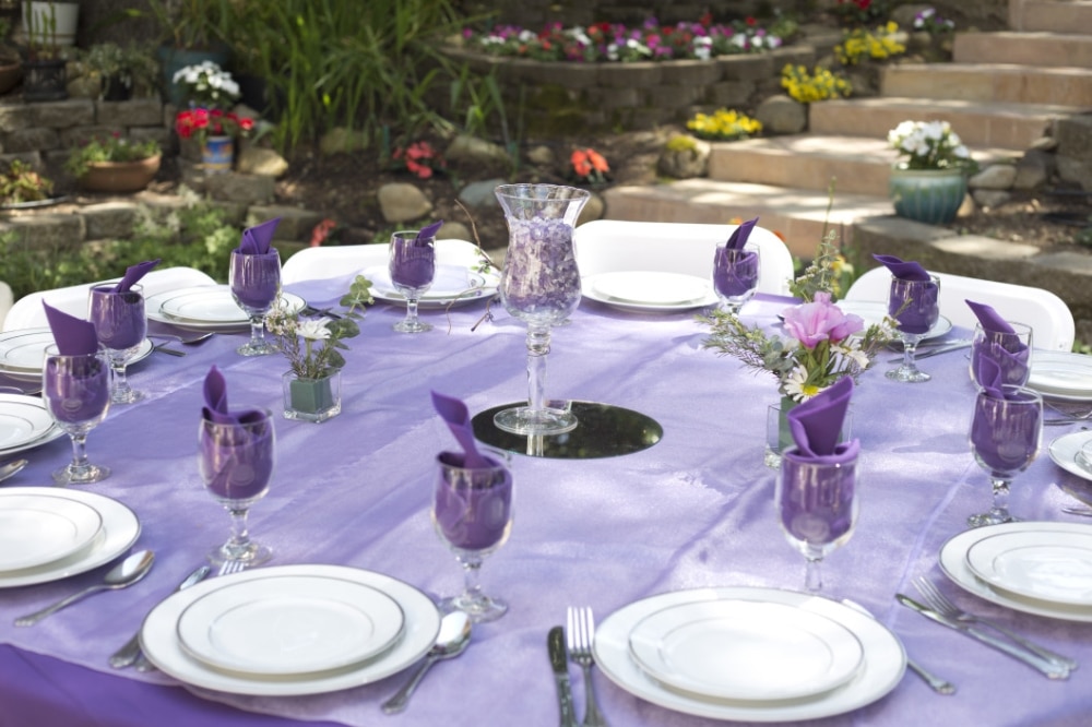 Purple and lavender wedding table setting with elegant glassware, floral centerpieces, and decorative napkins, surrounded by lush greenery and colorful flowers.