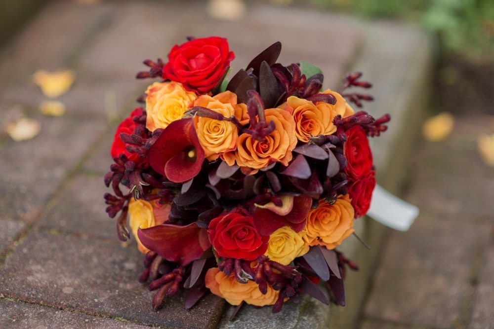 Wedding bouquet featuring vibrant red, orange, and yellow roses with dark foliage, set in a fall-inspired color scheme.