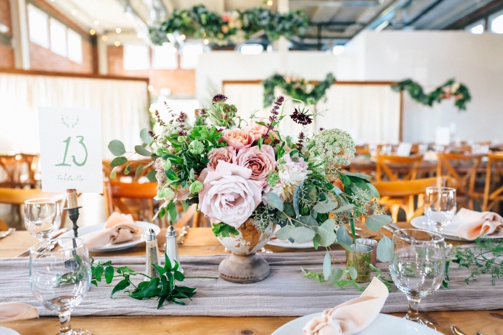 Elegant wedding table setting with a floral centerpiece in shades of pink and green, featuring table number 13, set against a rustic backdrop, enhancing a fall wedding theme with natural decor elements.
