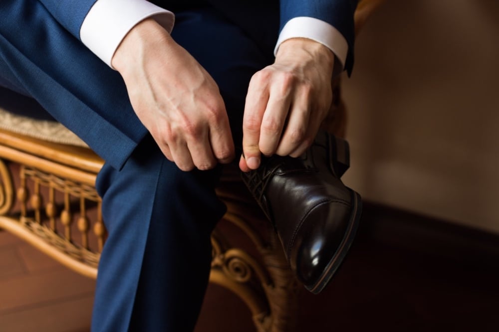 Groom adjusting dark brown dress shoes while preparing for a wedding, wearing a navy suit and white shirt.