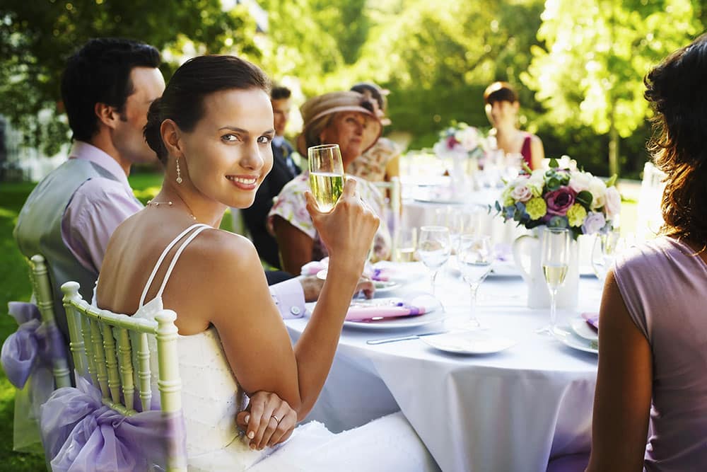 Bride smiling and holding a glass at wedding reception table surrounded by guests, with elegant decor and vibrant flowers, capturing a joyful moment of celebration.