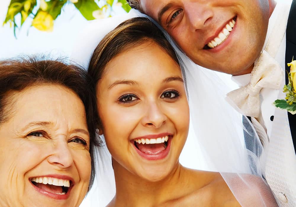 Happy bride and mother of the bride smiling together with groom, celebrating wedding joy in a vibrant outdoor setting.