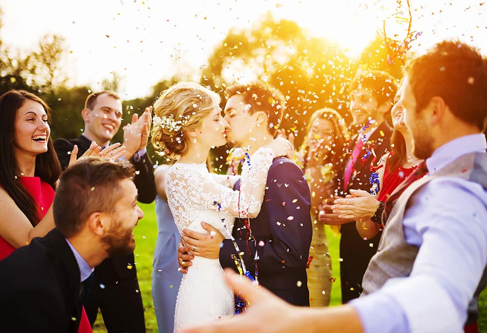 Newlywed couple kissing amidst confetti with joyful guests celebrating at a sunny outdoor wedding party.