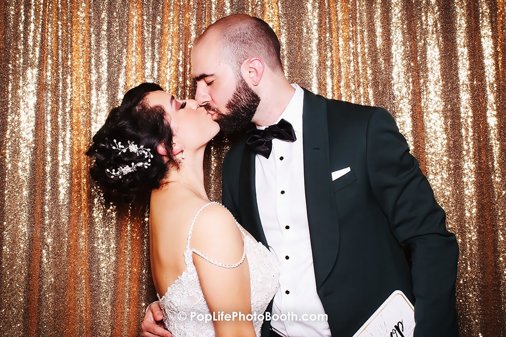 Newlywed couple kissing in photo booth with sparkling backdrop, capturing a moment of joy and celebration at their wedding gala.