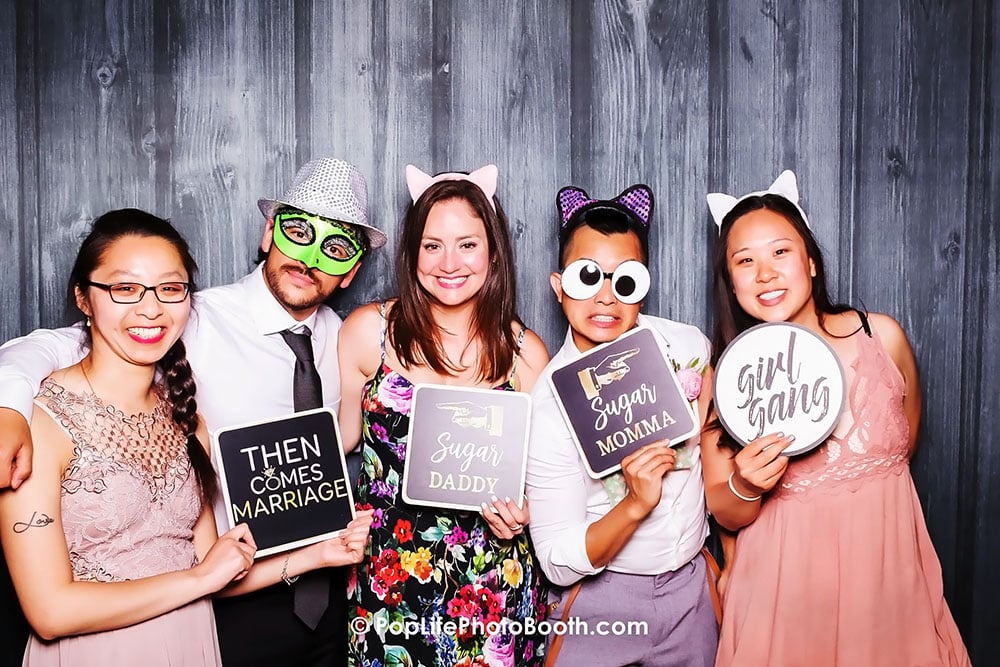 Group of friends having fun in a photo booth, holding playful signs like "Then Comes Marriage," "Sugar Daddy," "Sugar Momma," and "Girl Gang," against a stylish wooden backdrop, enhancing the wedding celebration atmosphere.