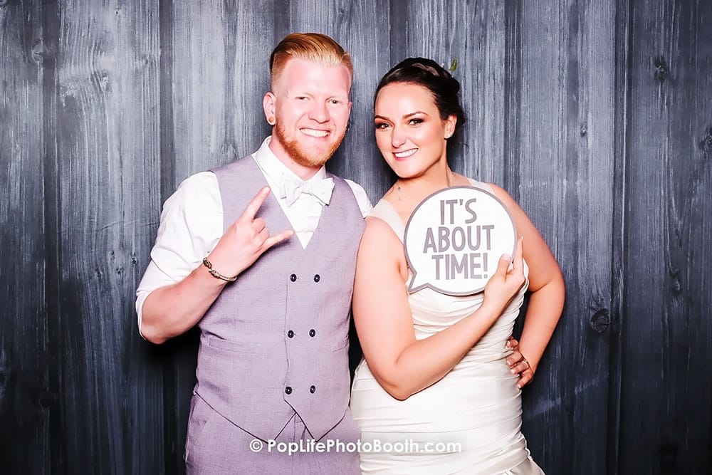 Happy newlywed couple posing in photo booth with sign reading "IT'S ABOUT TIME!" celebrating their wedding day.