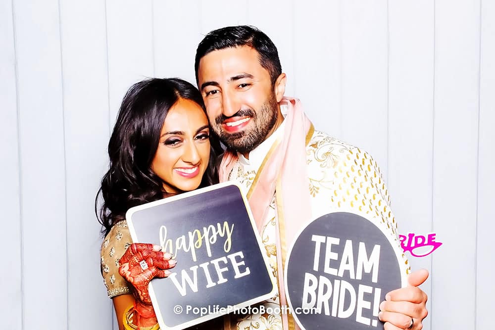 Happy couple celebrating in a photo booth, holding signs that say "happy WIFE" and "TEAM BRIDE," capturing joyful wedding memories.