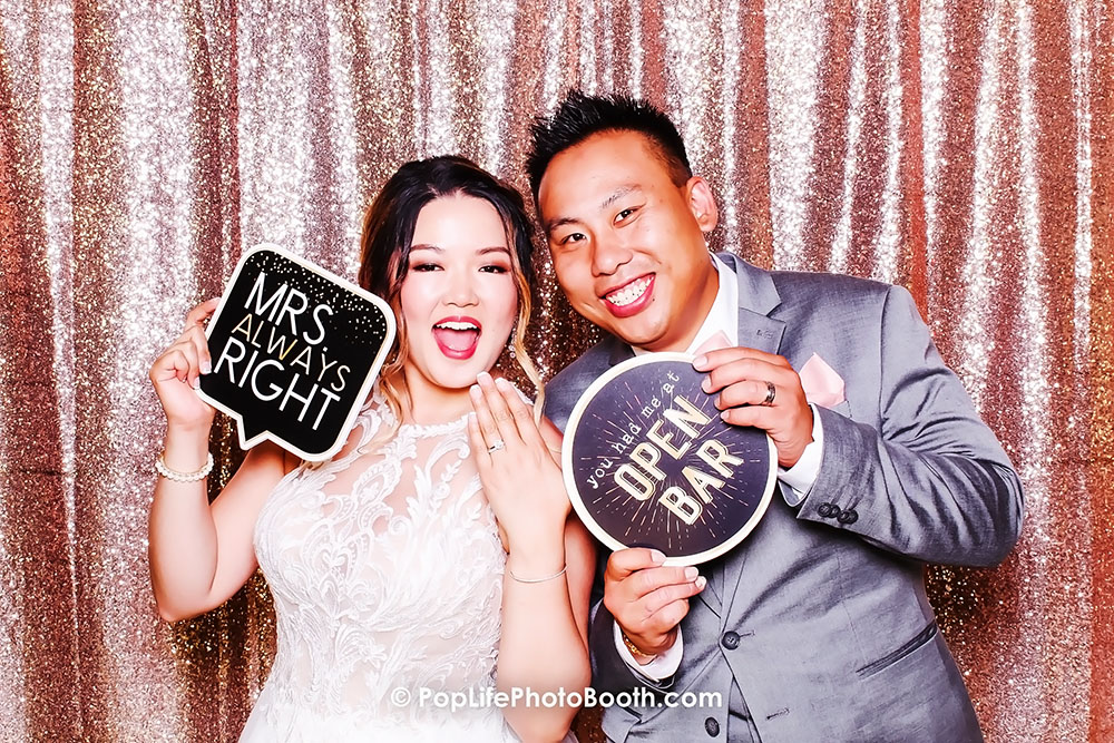 Newlywed couple posing in a photo booth, holding signs that read "MRS ALWAYS RIGHT" and "you had me at OPEN BAR," celebrating their wedding with joy and laughter against a sparkling backdrop.
