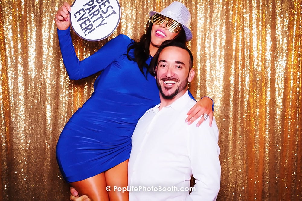 Happy guests posing in a photo booth, woman in a blue dress holding a sign reading "BEST PARTY EVER!" with a man smiling, gold sequin backdrop enhancing the festive atmosphere.